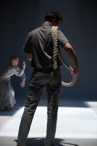 En man i gråa kläder med ett rep på sin axla, bakom en kvinna i en grå klänning dansar.